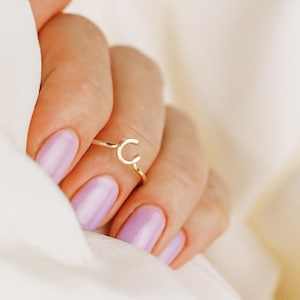 May include: A gold-colored ring featuring the letter "C" in a delicate, minimalist design. The ring is displayed on a hand with light purple nail polish, set against a soft, white background.