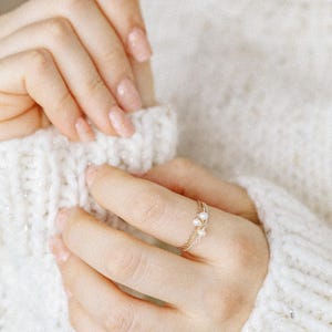 May include: A delicate gold ring featuring a thin, multi-strand band adorned with three small, white, oval-shaped freshwater pearls arranged in a cluster. The ring is crafted from fine gold wire, showcasing a minimalist and refined design.