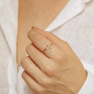 Peut inclure: Une délicate bague en fil d'or. La bague présente une forme de goutte d'eau avec un motif en boucle. La bague est portée au doigt, et l'arrière-plan est une chemise blanche.