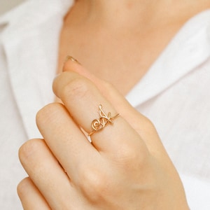Puede incluir: Un delicado anillo dorado con la letra cursiva "A". El anillo se lleva en un dedo, destacando su diseño elegante y minimalista. La construcción similar a un alambre del anillo añade un toque único a esta joya.