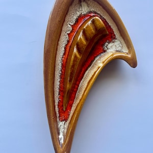 May include: A vintage ceramic ashtray in a unique, curved shape. The ashtray features a layered design with shades of brown, gold, red, and white. The interior has a deep red hue, while the exterior is a glossy brown.