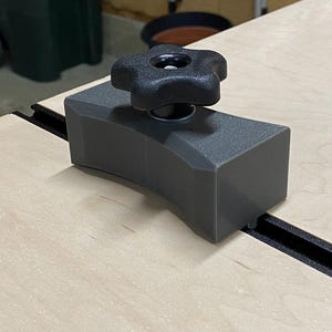 May include: A gray plastic woodworking clamp with a black knob. The clamp is attached to a black track on a wooden surface.