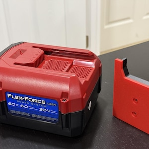 以下が含まれることがあります： 黒いラベルに「Flex-Force Power System L324 60 60 324」と書いてある赤い電動工具のバッテリー。バッテリーの隣には、2つの穴のある赤いプラスチック製のスタンドがある。