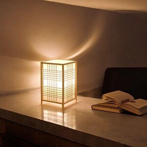 Pode incluir: Um abajur de mesa quadrado com um abajur branco e uma moldura de madeira. A lâmpada está em uma mesa de madeira com um livro em cima.