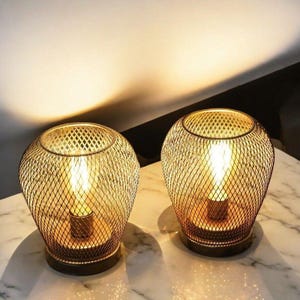 Pode incluir: Dois abajures de mesa de metal dourado com um design de malha de arame e uma lâmpada dentro. As lâmpadas estão sobre uma superfície branca.