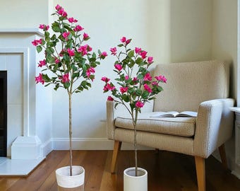Puede incluir: Dos árboles artificiales de buganvillas con flores rosas vibrantes y hojas verdes. Un árbol está en una cesta tejida, el otro en una maceta blanca. Los árboles están sobre un suelo de madera cerca de un sillón beige.
