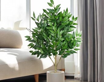 May include: A potted artificial plant with green leaves and a light gray pot. The plant is in a living room setting with a white couch and a window in the background.