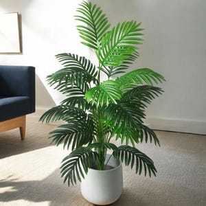 Puede incluir: Una gran palmera artificial verde en una maceta de cerámica blanca. La planta está en una habitación con una alfombra gris claro y un sofá azul.