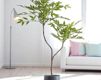 Könnte beinhalten: Ein künstlicher Baum mit grünen Blättern und einem braunen Stamm, gepflanzt in einem dunkelgrauen zylindrischen Topf. Der Baum steht in einem hellen Raum mit einem weißen Sofa und einer Stehlampe. Der Topf hat etwa 30 cm Durchmesser.