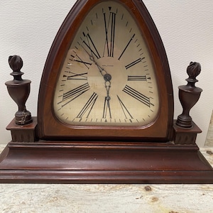 May include: A vintage wooden clock with a triangular face and Roman numerals. The clock has a dark brown finish and features the brand name "WALTHAM" on the clock face. Two decorative finials flank the clock face.