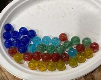 May include: A collection of colorful glass marbles arranged in a white, round dish. The marbles are in shades of blue, teal, green, red, and yellow. The marbles have a swirled pattern. The dish is on a brown surface.