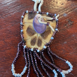 May include: A beaded turtle shell pendant with a purple shell inside and a white leather cord. The pendant has a fringe of purple and white beads.