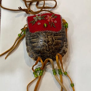 May include: A small, red, beaded pouch with a turtle shell bottom. The pouch is decorated with flowers and beads. It is attached to a brown leather cord.