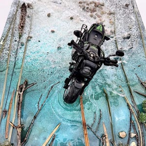May include: A miniature black motorcycle with green accents is positioned on a textured, mixed-media art piece. The artwork features a teal and white color scheme, with embedded natural elements like twigs and stones, creating a landscape scene.