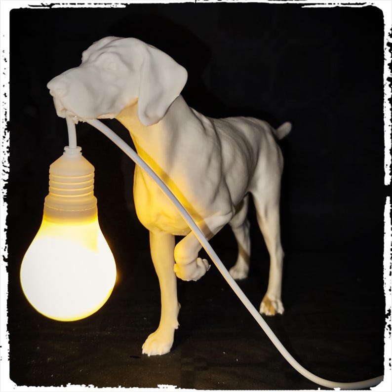 以下が含まれることがあります： 口に電球を持った白いセラミック製の犬の彫刻。犬は黒い表面に立っており、電球は点灯しています。