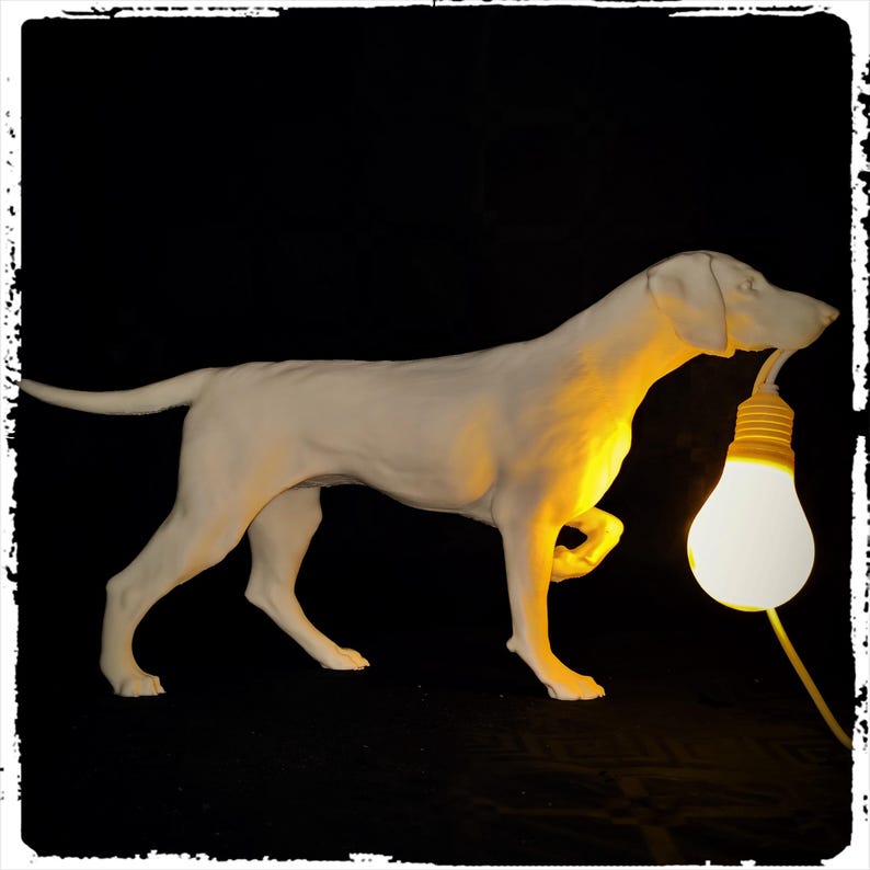 以下が含まれることがあります： 犬が電球を持っている白いセラミックの彫刻。電球は点灯しており、黄色く光っています。