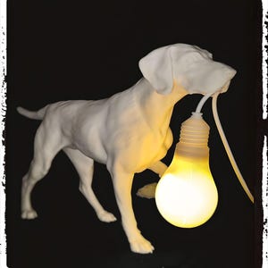 Könnte beinhalten: Weiße Keramik-Hundeskulptur, die eine leuchtende Glühbirne hält. Der Hund steht auf einer schwarzen Oberfläche.