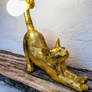 Può includere: Lampada a forma di gatto color oro in posizione allungata. La lampada ha una lampadina alla fine del collo. È posizionata su una superficie di legno. Il corpo del gatto ha una finitura metallica strutturata.