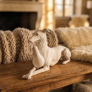 Può includere: Una scultura in bianco opaco di un levriero in posizione sdraiata, che indossa un collare e tiene una lampadina bianca in bocca. La scultura è realizzata con superfici lisce e solide, presentando dettagli anatomici realistici del cane.
