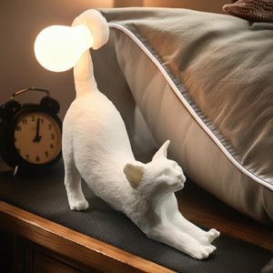 Könnte beinhalten: Eine weiße katzenförmige Lampe mit einer Glühbirne als Kopf, die sich nach vorne streckt. Die Lampe steht auf einer Holzoberfläche, mit einem schwarzen Wecker und einem Kissen im Hintergrund. Die Glühbirne der Lampe leuchtet.