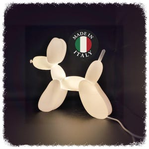 Puede incluir: Una lámpara de perro globo blanco e iluminado. La lámpara tiene la forma de un perro hecho de globos conectados. La imagen incluye una pegatina "Made in Italy" con la bandera italiana. La lámpara está sobre una superficie oscura.