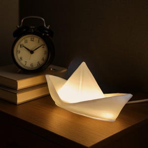 Könnte beinhalten: Ein weißes, papierbootförmiges Nachtlicht mit warmem, weißem Licht. Das Licht steht auf einer Holzoberfläche neben einem schwarzen Wecker.