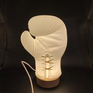 Puede incluir: Una lámpara con forma de guante de boxeo blanco con una base de madera. La lámpara está encendida y el cable es visible.