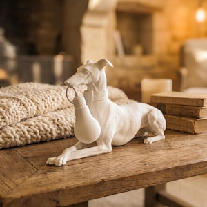 Può includere: Una statuetta in ceramica bianca raffigurante un levriero in posizione sdraiata. Il cane indossa un collare e tiene una lampadina bianca in bocca. La scultura presenta una finitura opaca liscia e dettagli anatomici della forma canina.