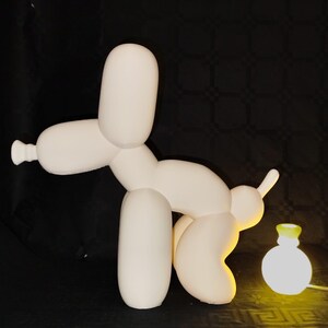 May include: White ceramic sculpture of a balloon dog in a playful pose. The dog is standing on a black surface and is illuminated by a yellow light.