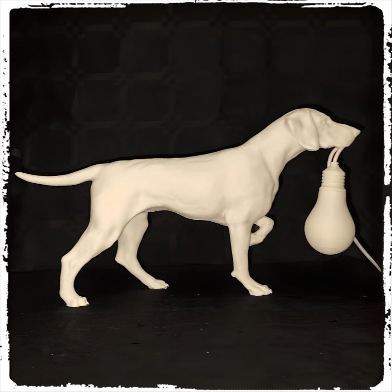 以下が含まれることがあります： 犬が電球を持っている白いセラミック製の彫刻。犬は四つん這いで立っており、頭を横に傾けている。電球は犬の口からぶら下がっている。