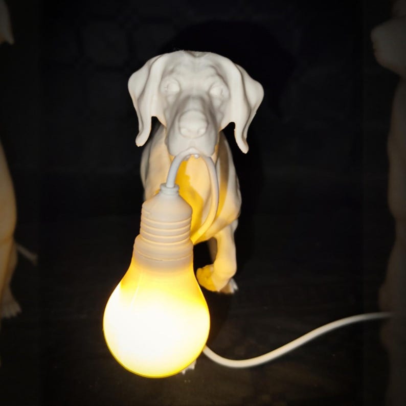 以下が含まれることがあります： 白いセラミック製の犬の彫刻が電球を持っています。電球は点灯しており、黄色く光っています。