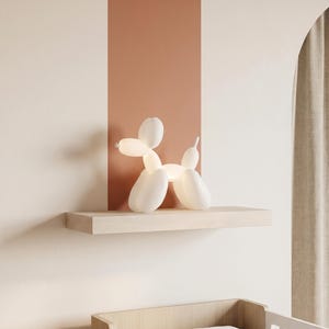Op de afbeelding: Een witte ballonhond-lamp die een zachte, diffuse gloed uitstraalt. De lamp heeft de vorm van een gestileerd dier gemaakt van gedraaide ballonnen, met een ronde kop, een langwerpige snuit, vier poten en een kleine staart, in een matte afwerking.