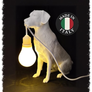 Op de afbeelding: Een witte, hondvormige lamp met een matte afwerking, afgebeeld in een zittende houding. De hond houdt een gloeiende, warmgekleurde lamp in de bek, die is verbonden met een wit snoer. Een rond embleem bevat de tekst Made in Italy.
