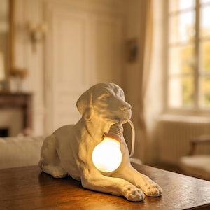 Op de afbeelding: Een witte keramische tafellamp in de vorm van een liggende hond die een gloeiende ronde gloeilamp in zijn bek houdt. De lamp heeft een gladde matte afwerking en een wit stroomsnoer dat uit de fitting komt, rustend op een houten oppervlak.