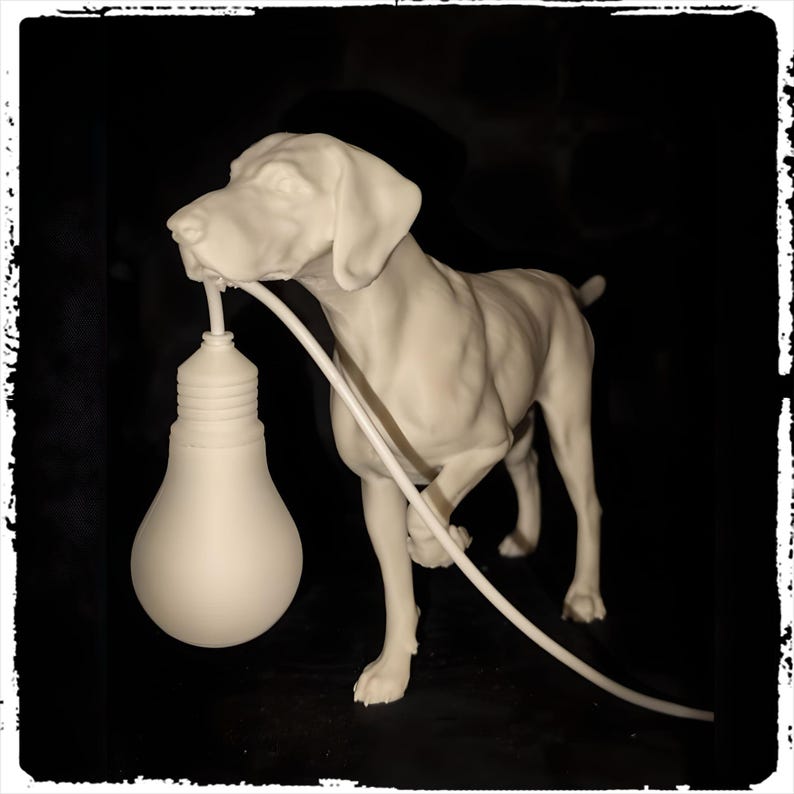 以下が含まれることがあります： 口に大きな電球を持った犬の白いセラミック彫刻。犬は黒い表面に立っており、電球は白いコードにつながっています。