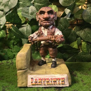 May include: A detailed figurine of a goblin-like character from the film Labyrinth, holding a staff. The figure is dressed in brown and tan clothing and stands on a platform with the word "LABYRINTH" on it. The background features green foliage.
