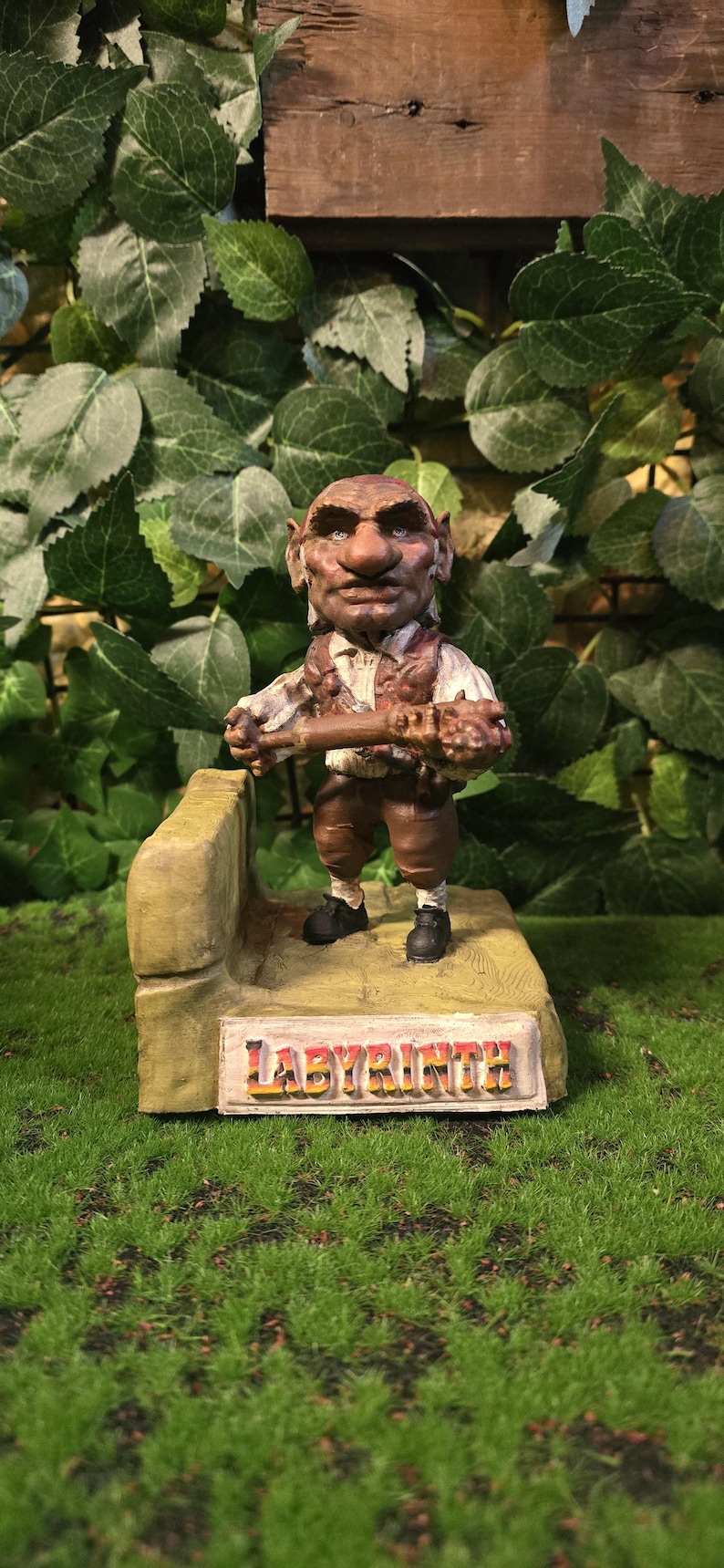 May include: A detailed figurine of a goblin-like character from the film Labyrinth, holding a staff. The figure is dressed in brown and tan clothing and stands on a platform with the word "LABYRINTH" on it. The background features green foliage.