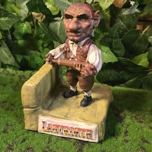 May include: A detailed figurine of a goblin-like character from the film Labyrinth, holding a wooden instrument. The figure is brown with a red cap and vest, standing on a stone base with the word "LABYRINTH" on it.