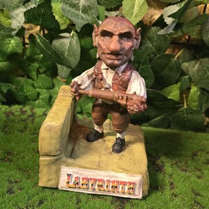 May include: A detailed figurine of a character from the film Labyrinth. The figure is brown and tan, holding a club, and standing on a base with the word "Labyrinth" on it. The background is green.