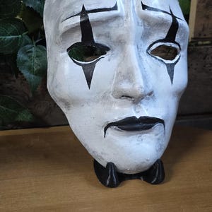 May include: A white mask with black accents, depicting a stylized face with open eye sockets and a black bow tie. The mask has black markings above the eyes and on the cheeks. The mask is on a wooden surface.