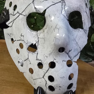 May include: A white hockey mask with a cracked and weathered appearance. The mask has a large eye hole and several smaller holes throughout. The mask is on a black stand.