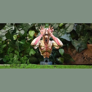 Puede incluir: Busto detallado de una criatura con costillas expuestas, piel pálida y dedos largos en forma de garra. Los brazos de la figura están levantados y su rostro está contorsionado en un grito. El busto está montado sobre una base azul oscuro, sobre un fondo de follaje verde.