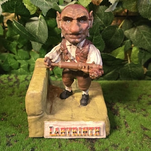 Op de afbeelding: Gedetailleerd beeldje van een goblinachtig personage uit de film Labyrinth. De figuur is bruin en bruin, met een houten voorwerp. De basis heeft het woord "LABYRINTH" in gouden letters. De achtergrond heeft groen gebladerte.