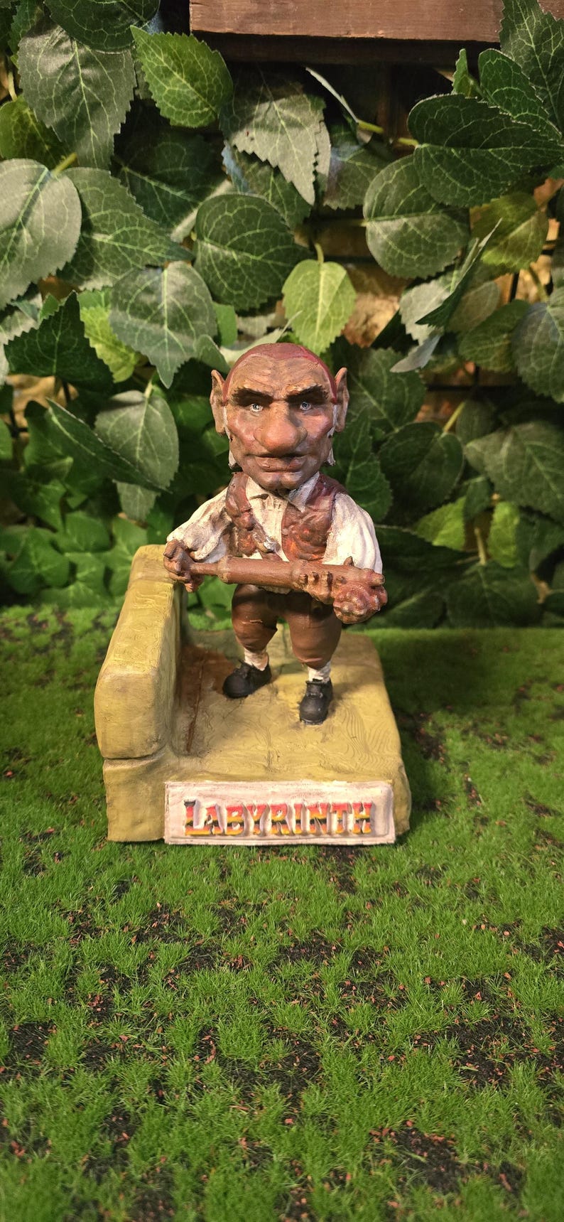 May include: A detailed figurine of a goblin-like character from the film Labyrinth. The figure is brown and tan, holding a wooden object. The base has the word "LABYRINTH" in gold lettering. The background features green foliage.