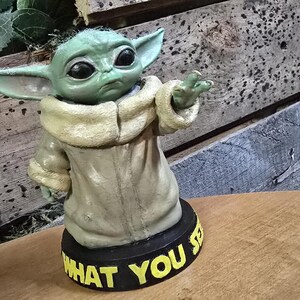 Peut inclure: Une figurine verte et brune d'un personnage de la franchise Star Wars, connu sous le nom de "Baby Yoda" ou "L'Enfant". La figurine est posée sur une base noire avec les mots "What You See" écrits en lettres jaunes.