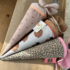 Könnte beinhalten: Drei Stoffkegel mit verschiedenen Mustern. Der erste Kegel ist rosa mit braunen Herzen, der zweite Kegel ist hellgrau mit braunen Kreisen und der dritte Kegel ist braun mit Leopardenmuster. Die Kegel sind mit einem rosa Band gebunden.