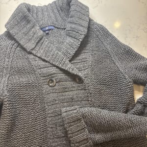 May include: A gray knit cardigan with a shawl collar and two large buttons. The sweater has a double-breasted design and a textured knit pattern. The Gap brand label is visible on the back of the collar.
