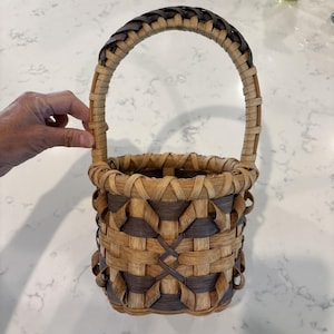 May include: A hand-woven basket with a rounded handle. The basket features a combination of natural and dark brown woven materials, creating a geometric pattern on the sides. The handle is a braided design, and the basket is designed for carrying items.