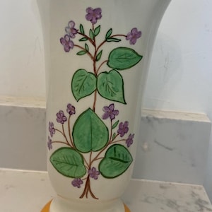 May include: A white ceramic vase with a flared top and a round base, both trimmed in orange. The vase is decorated with a hand-painted floral design featuring green leaves and purple flowers. A decorative piece for the home.