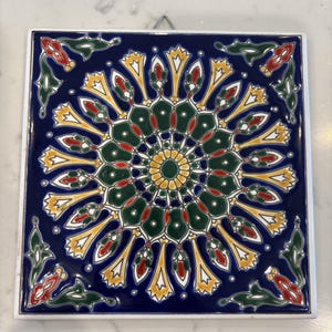 May include: Square ceramic tile with a detailed floral design. The tile features a dark blue background with a central circular pattern in yellow, green, and red. The corners have additional floral accents.